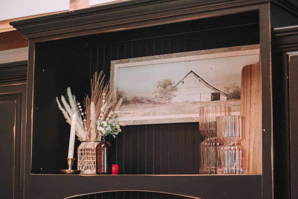 A dark wood hutch holding a framed painting of a barn, dried flowers, and glassware