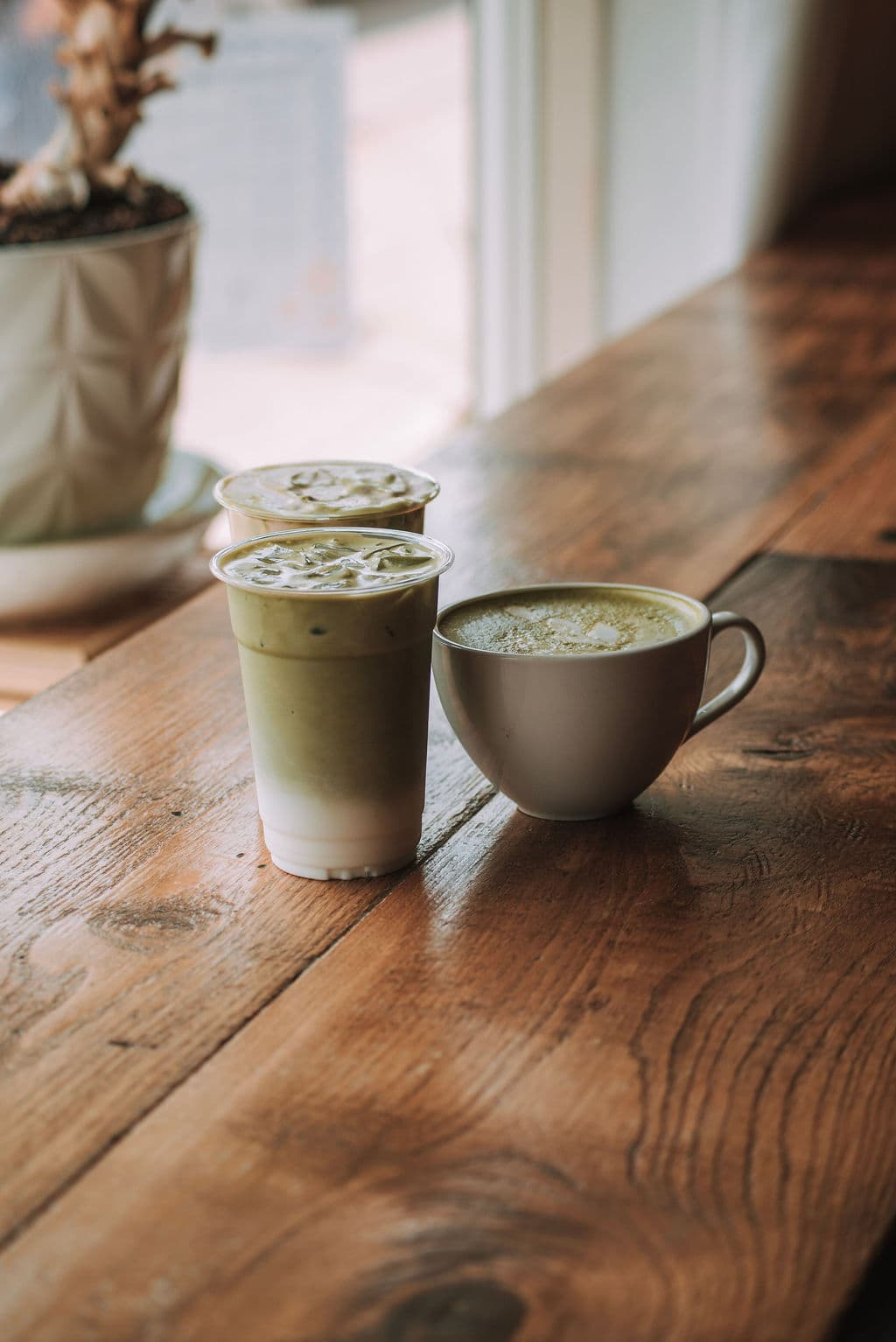 An iced matcha latte and a cappuccino on a reclaimed wood table in the window light