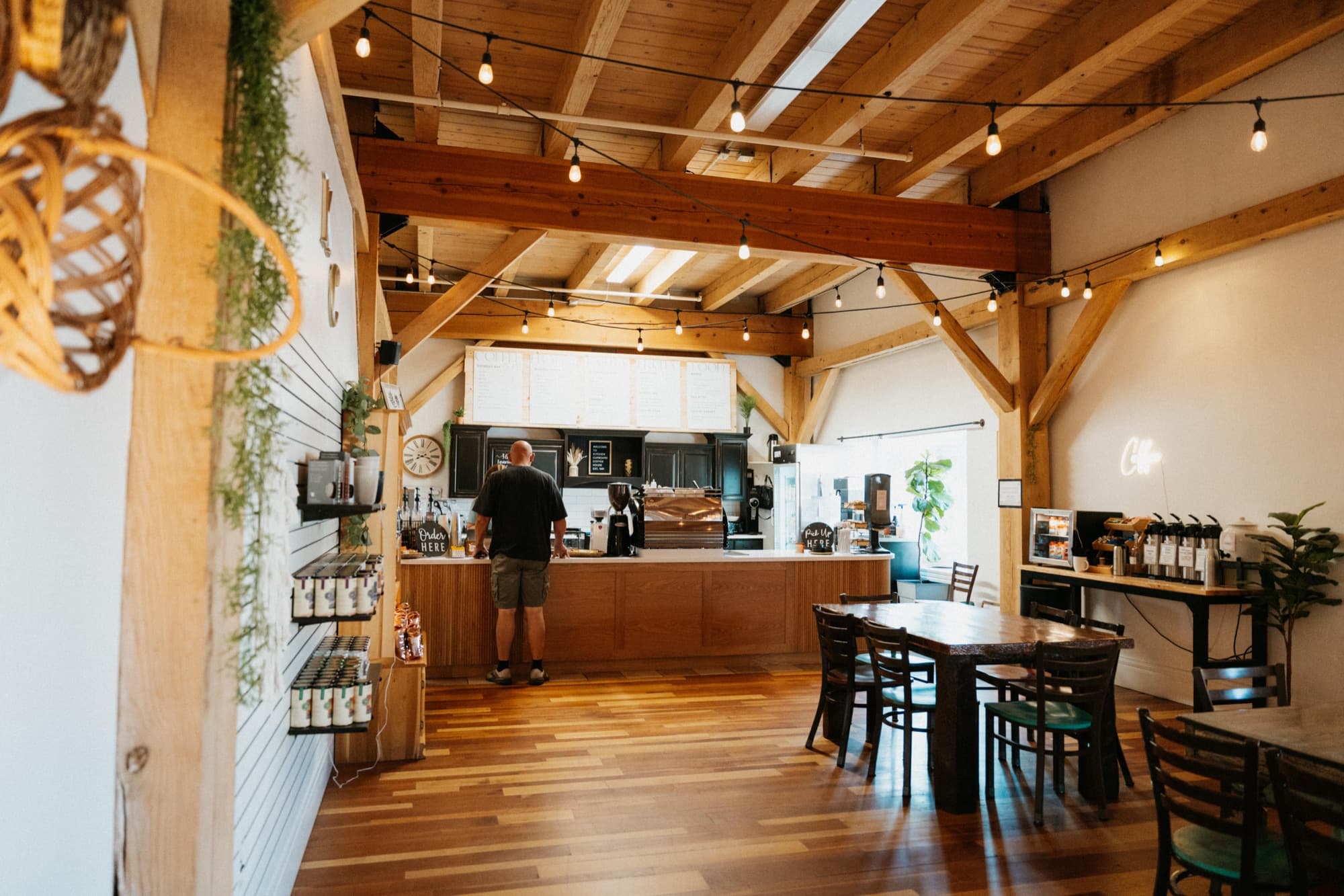 The dining room under exposed timber trusses and string lights, looking toward the service counter, with retail shelves along the left wall