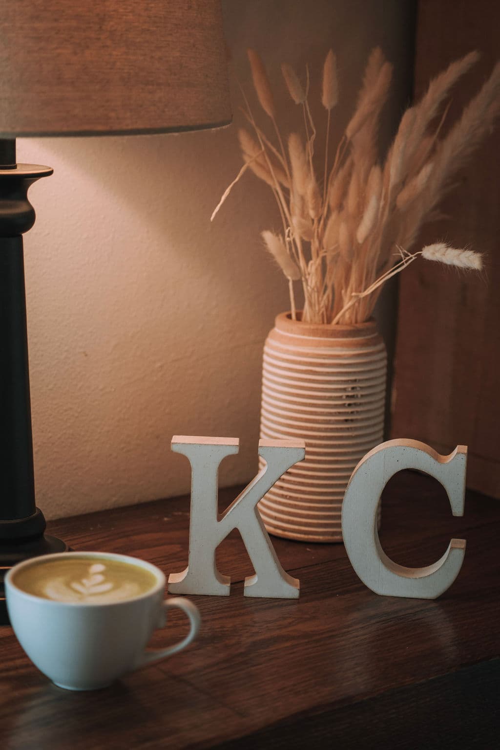 The wooden KC monogram letters standing beside a latte and dried wheat